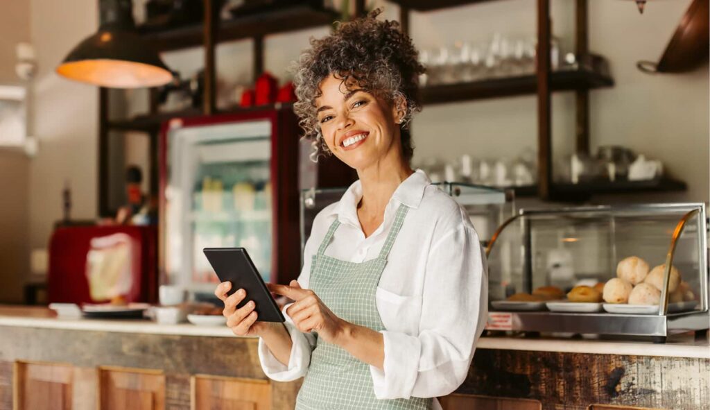 Image of a female café owner in her venue smiling at the camera and holding an ipad.