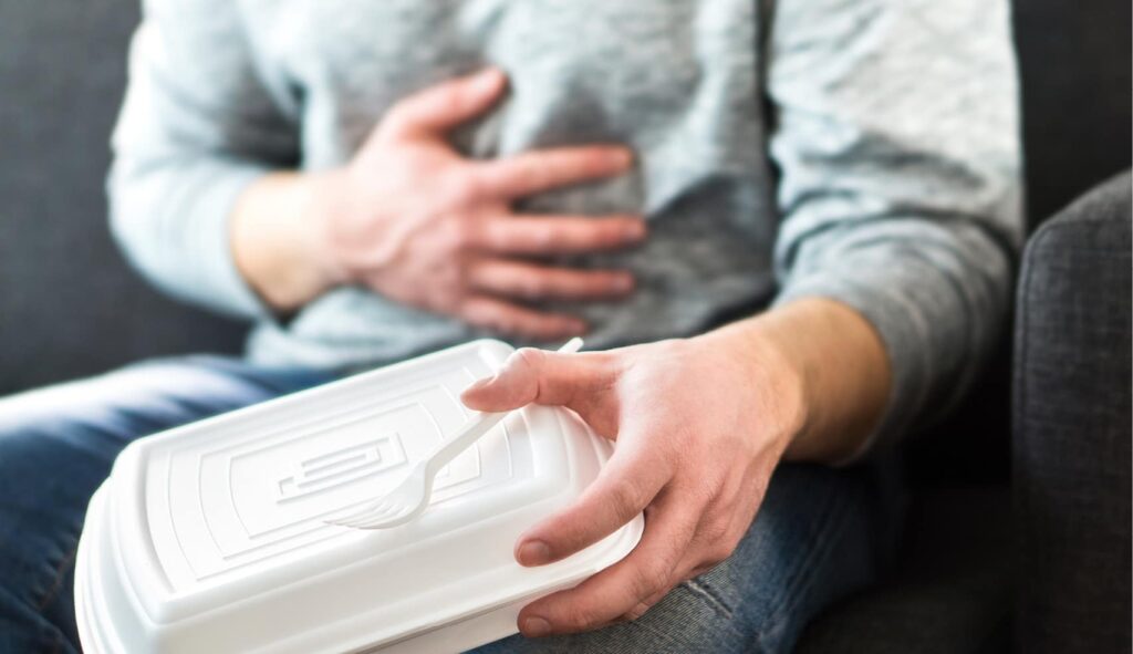 An image of a person experiencing food poisoning with hand on tummy and other hand holding a takeaway container. 